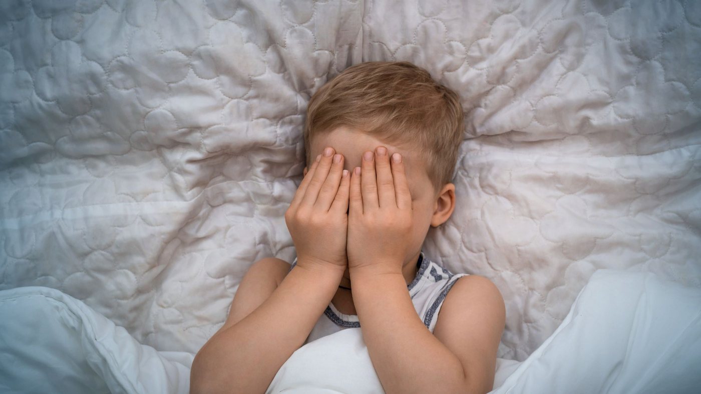 Overprikkeld hoogsensitief jongetje ligt in bed en houdt zijn handen voor zijn ogen
