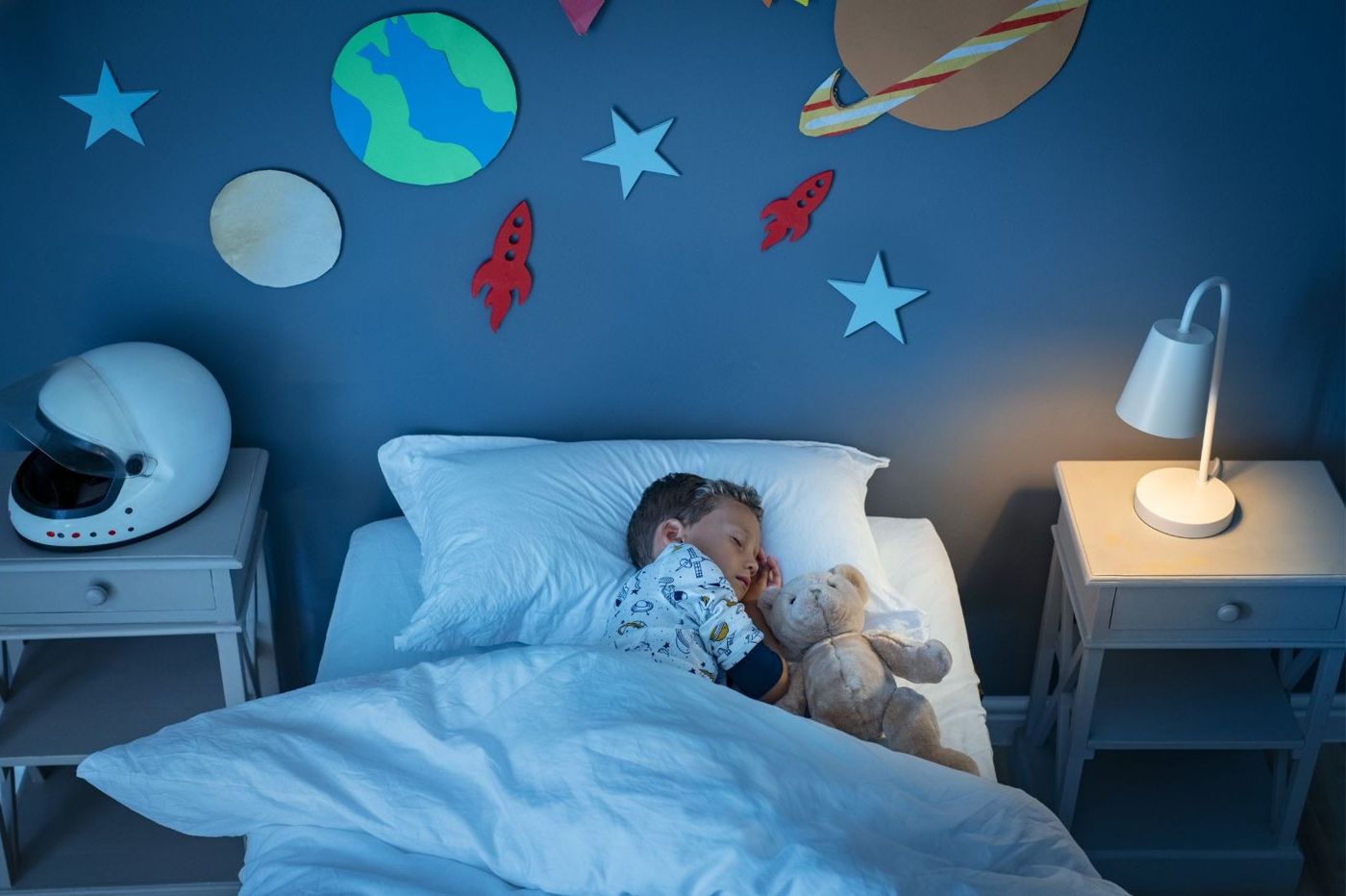 Jongen ligt met zijn knuffel in bed in een donkere kinderkamer met alleen een klein lampje aan