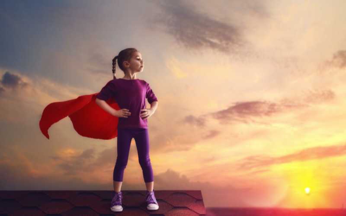 Meisje met superheldencape staat sterk en zelfverzekerd op een dak in de avondzon