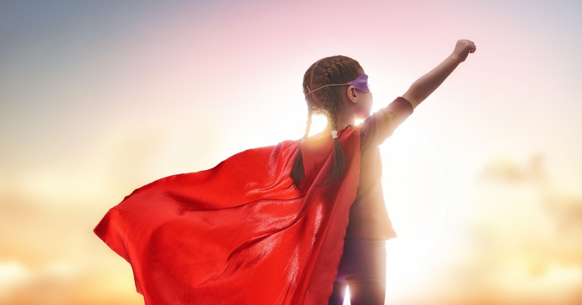 Kind met superheldencape juicht met de armen omhoog voor de ondergaande zon